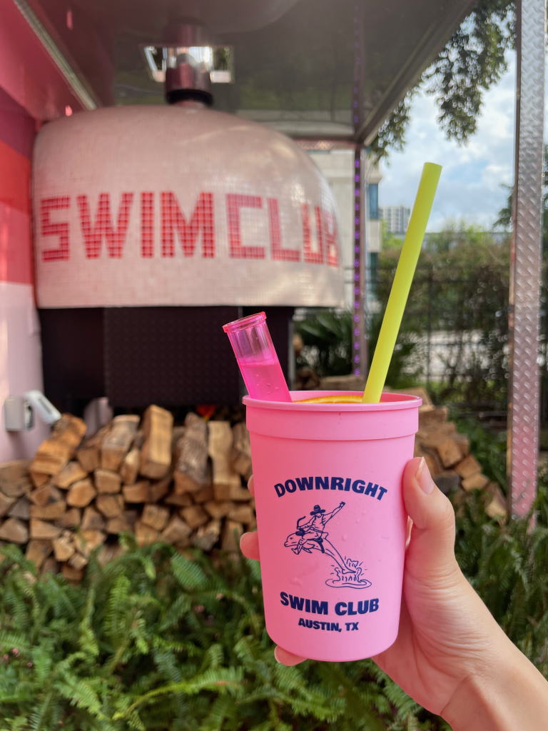 Pink cup that says "Downright Austin, Swim Club Austin, TX" with a cowboy riding a dolphin graphic on it. Behind it is a wood fired oven for pizzas with pink and white tiling that spells "Swim Club" on it, as well as some firewood and ferns just below it.